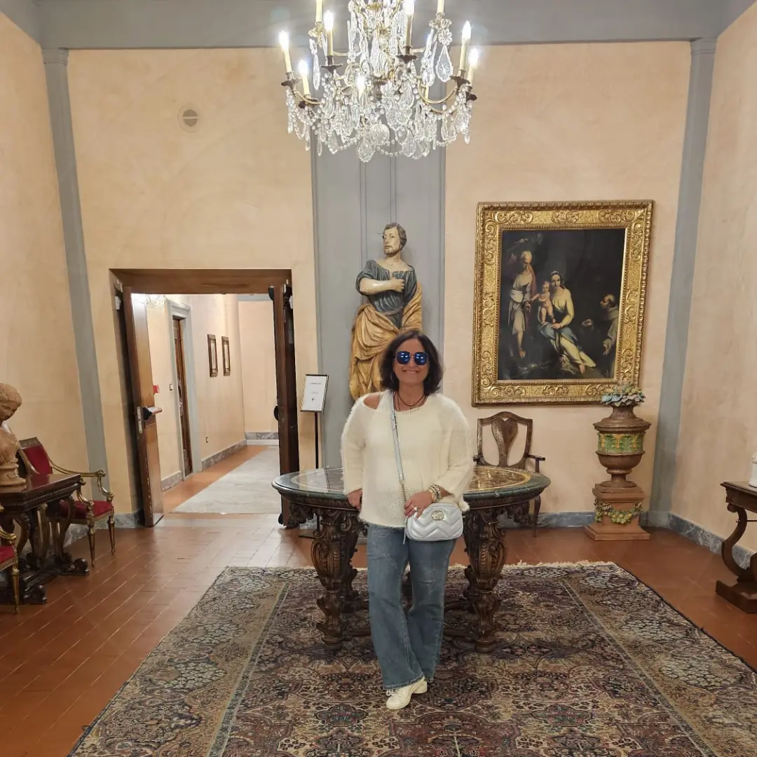 A luxurious Italian interior with a polished wooden floor and an ornate Persian rug. A woman stands in the center, wearing a white sweater, jeans, and sunglasses, holding a white handbag. The room features a grand chandelier, a carved wooden table, a gold-framed painting, a classical statue, and elegant furniture with red upholstery, reflecting a rich cultural setting for Italian Speaking Classes inspiration.